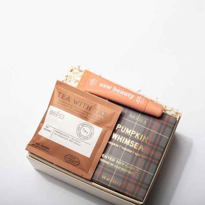 A curated gift box set on a clean white background, featuring a tea packet labeled 'TEA WITH ME', a peach-colored lip balm tube from 'ear beauty', and a plaid book or booklet titled 'PUMPKIN WHISPER'.