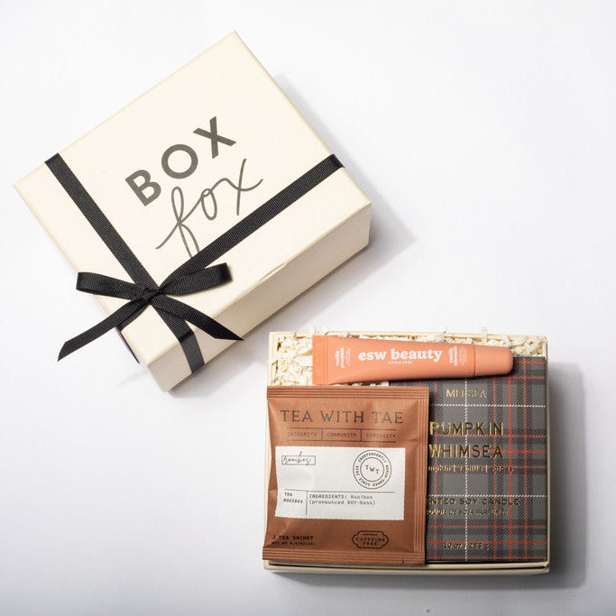 A curated gift box set from Box Fox, featuring a cream-colored gift box with a black ribbon and bow alongside tea and beauty items including a Tea With Tae packet and a pink New Beauty stick.