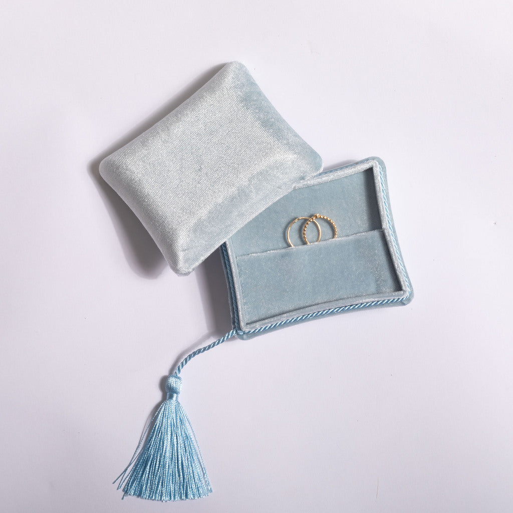 A light blue velvet ring pouch with a matching tassel, shown partially open with two gold rings inside on a plain light background.