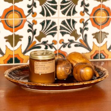 An orange candle with a gold lid with the description of, &quot; NO. 01: SPICED PUMPKIN SOY CANDLE,&quot; poured in California with Soy Wax, sitting on a brown bowl on a table. There is a floral background wall.