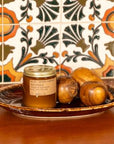 An orange candle with a gold lid with the description of, " NO. 01: SPICED PUMPKIN SOY CANDLE," poured in California with Soy Wax, sitting on a brown bowl on a table. There is a floral background wall.