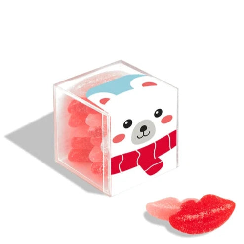 A small transparent cube-shaped box filled with red and pink sugar coated candies shaped like lips. The box is decorated with a cute polar bear wearing a red scarf. Two candies are placed outside the box for display.