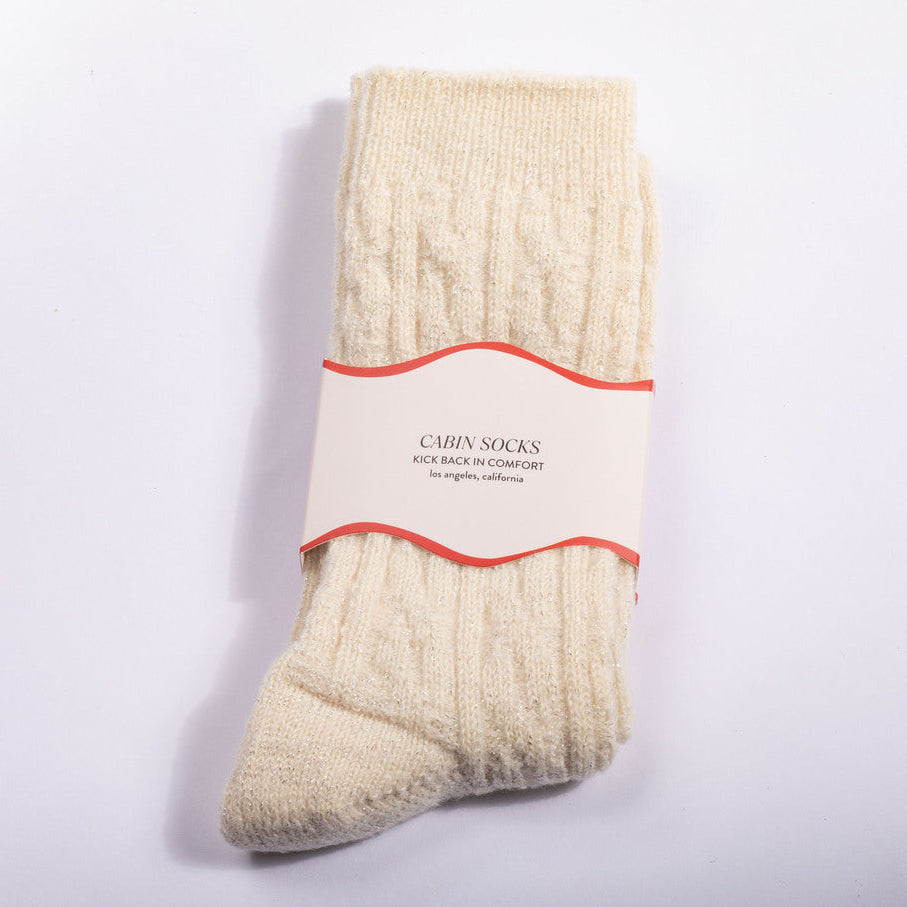 A pair of cream-colored cable-knit socks wrapped in a paper band label that reads 'CABIN SOCKS KICK BACK IN COMFORTABLE size options available'.