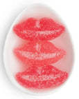 A clear oval dish holding three reddish-pink sugar coated gummy candies shaped like lips, displayed on a white background. 