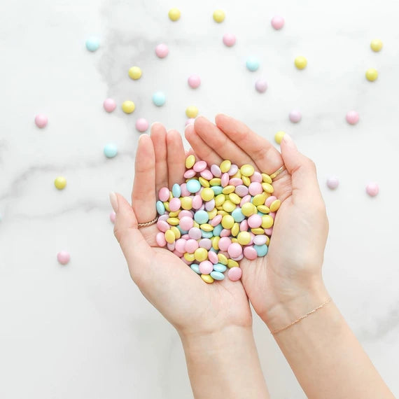 hands holding pink, purple, blue, and yellow coated chocolate pieces 