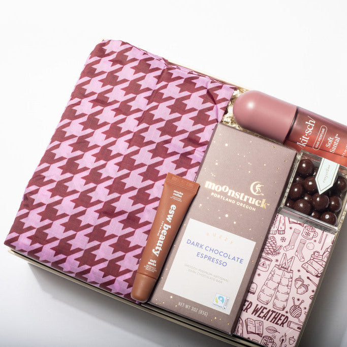 A curated gift box set on a white background, featuring a pink and burgundy houndstooth pouch, a box of Moonstruck Dark Chocolate Espresso, an Avery soap, and a Weatherproof pen in a clear plastic tray.