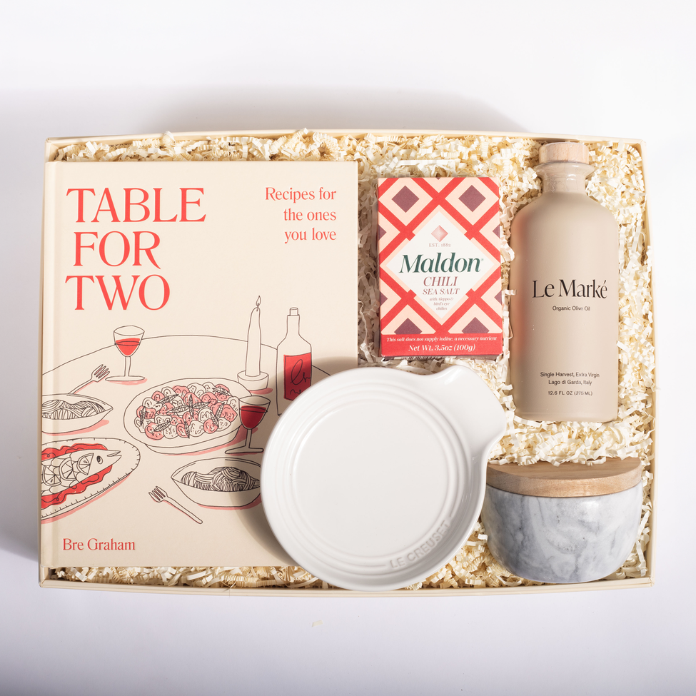 A curated gift box set on a white background, containing a book titled 'Table for Two' by Bre Graham, a box of Maldon Cheddar Cheese, a bottle of Le Marké Organic Olive Oil, a white ceramic bowl, and a marble-patterned jar with a wooden lid, all arranged in a kraft paper shred box.