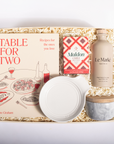 A curated gift box set on a white background, containing a book titled 'Table for Two' by Bre Graham, a box of Maldon Cheddar Cheese, a bottle of Le Marké Organic Olive Oil, a white ceramic bowl, and a marble-patterned jar with a wooden lid, all arranged in a kraft paper shred box.