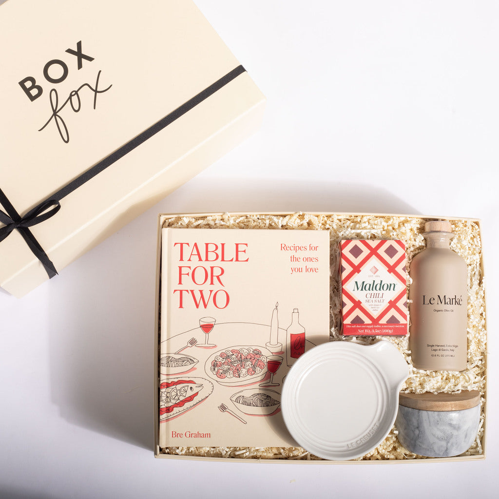 A curated gift box set from Box Fox, featuring the book 'Table for Two' by Bee Graham, a bottle of Le Marnier, a small white bowl, and a marble candle tin, arranged in a kraft paper box with shredded paper filler.