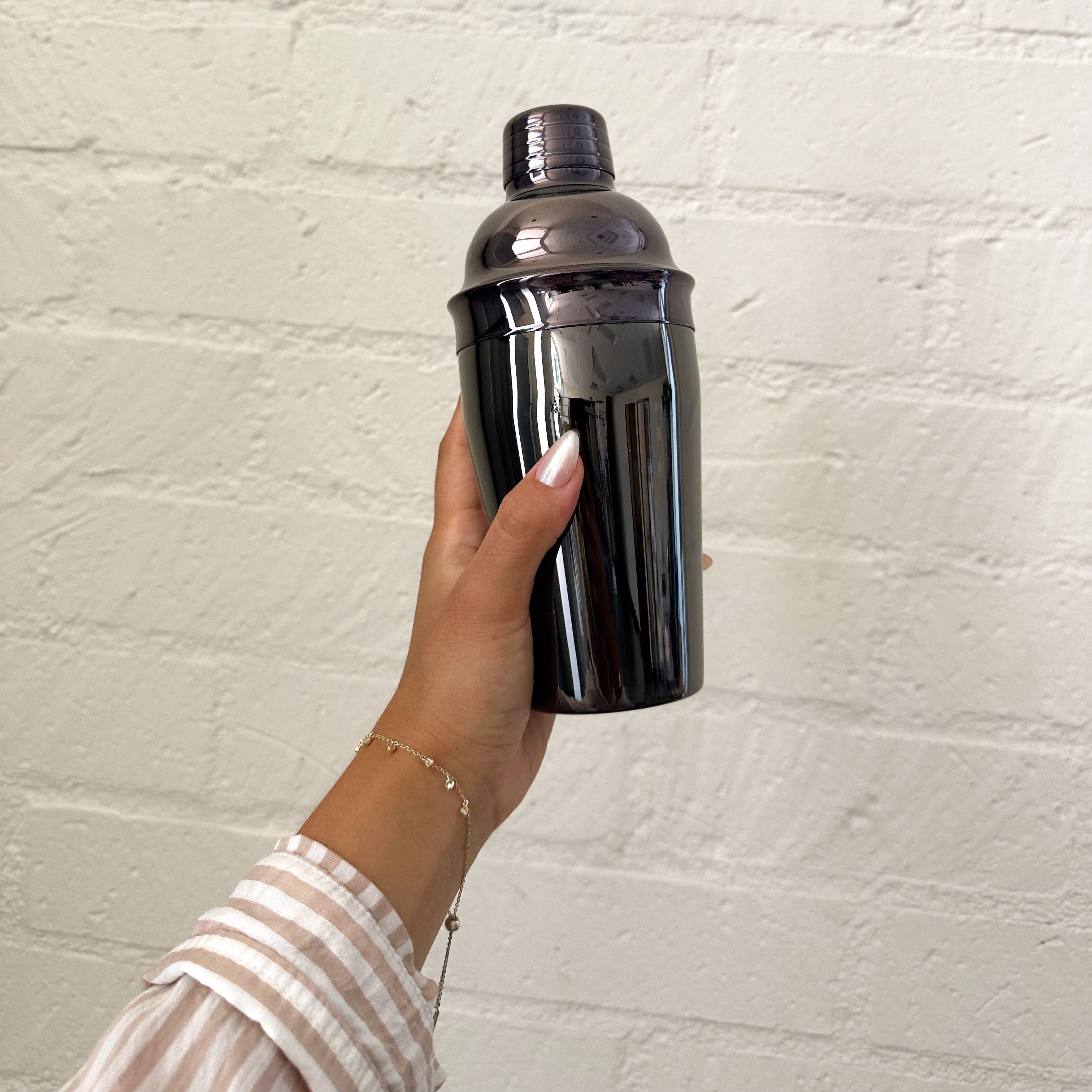 Person holding a black cocktail shaker against a light brick wall.