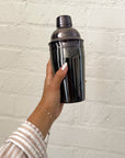 Person holding a black cocktail shaker against a light brick wall.