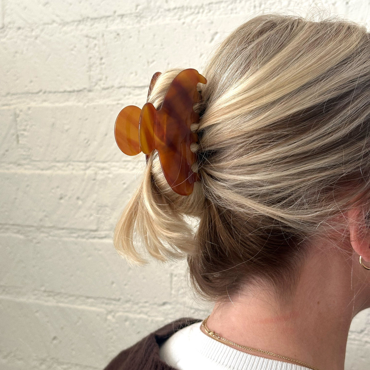 Person with blonde hair tied back using a tortoiseshell hair clip against a white brick wall.