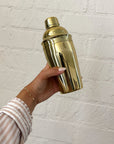 Gold cocktail shaker held by a hand against a white brick wall.