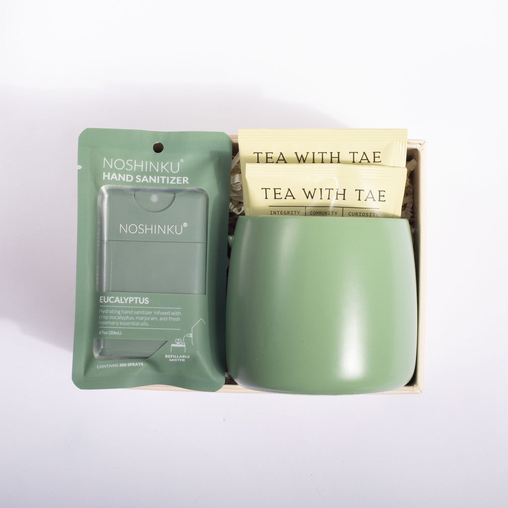 A gift set containing a green Noshinku hand sanitizer packet and a matching sage green ceramic mug filled with Tea With Tae tea bags, presented in a light wood box on a white background.