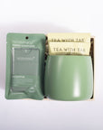 A gift set containing a green Noshinku hand sanitizer packet and a matching sage green ceramic mug filled with Tea With Tae tea bags, presented in a light wood box on a white background.