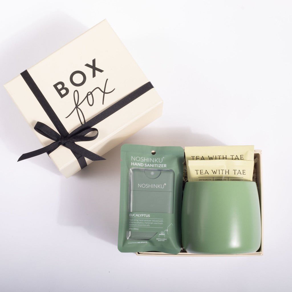 A gift set on a white background featuring a cream-colored 'BOX fox' gift box with a black ribbon, alongside an open package containing a sage green Noshinklu hand sanitizer packet and a matching sage green ceramic cup with two tea bags labeled 'TEA WITH TAE'.