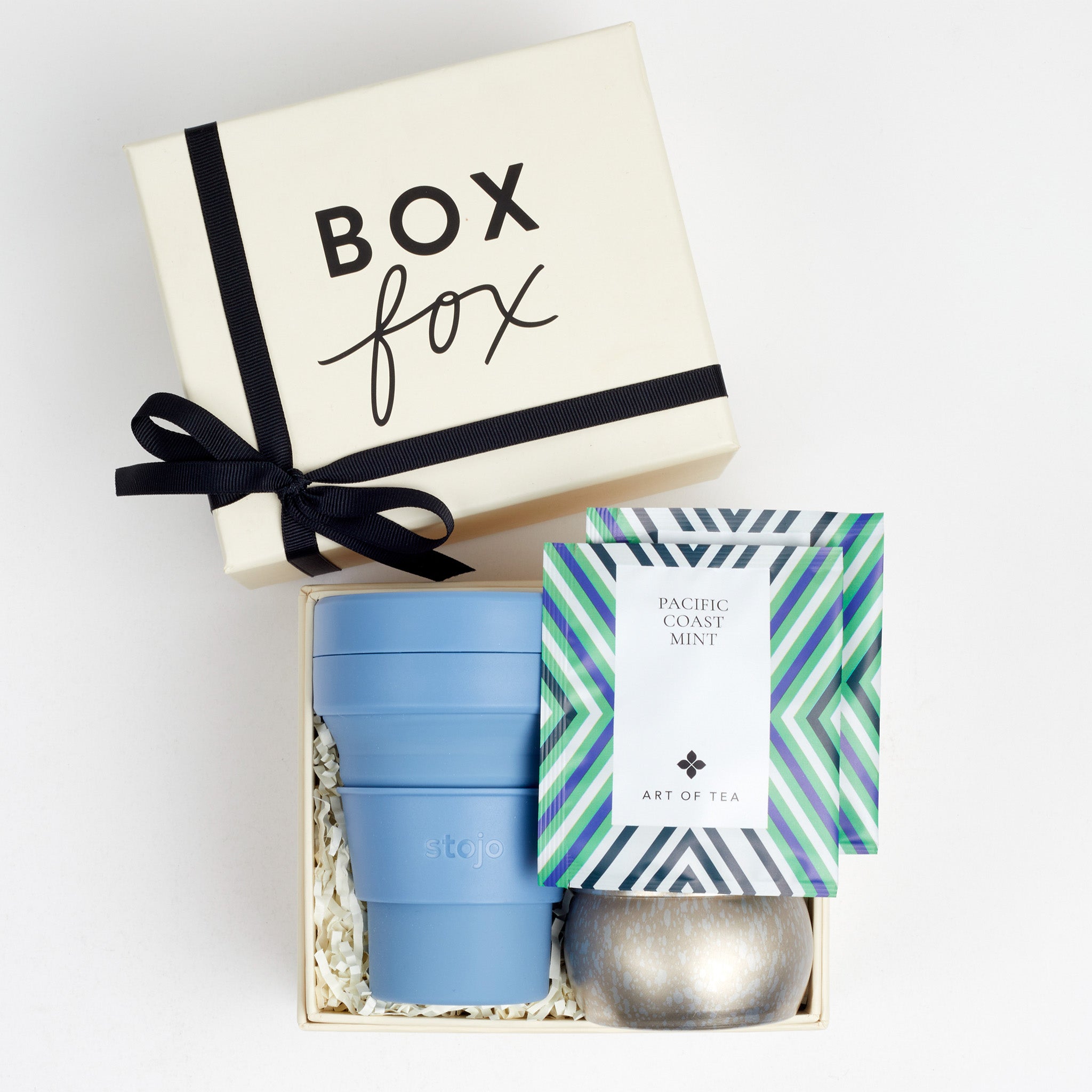 Creme BOXFOX gift box with a bow, next to an open gift box containing a blue collapsible cup, a silver tin candle and two art of tea sachets.