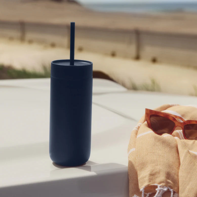 Navy blue tumbler with a straw on a ledge with sunglasses and a towel in the background.