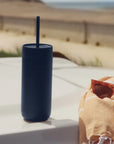Navy blue tumbler with a straw on a ledge with sunglasses and a towel in the background.