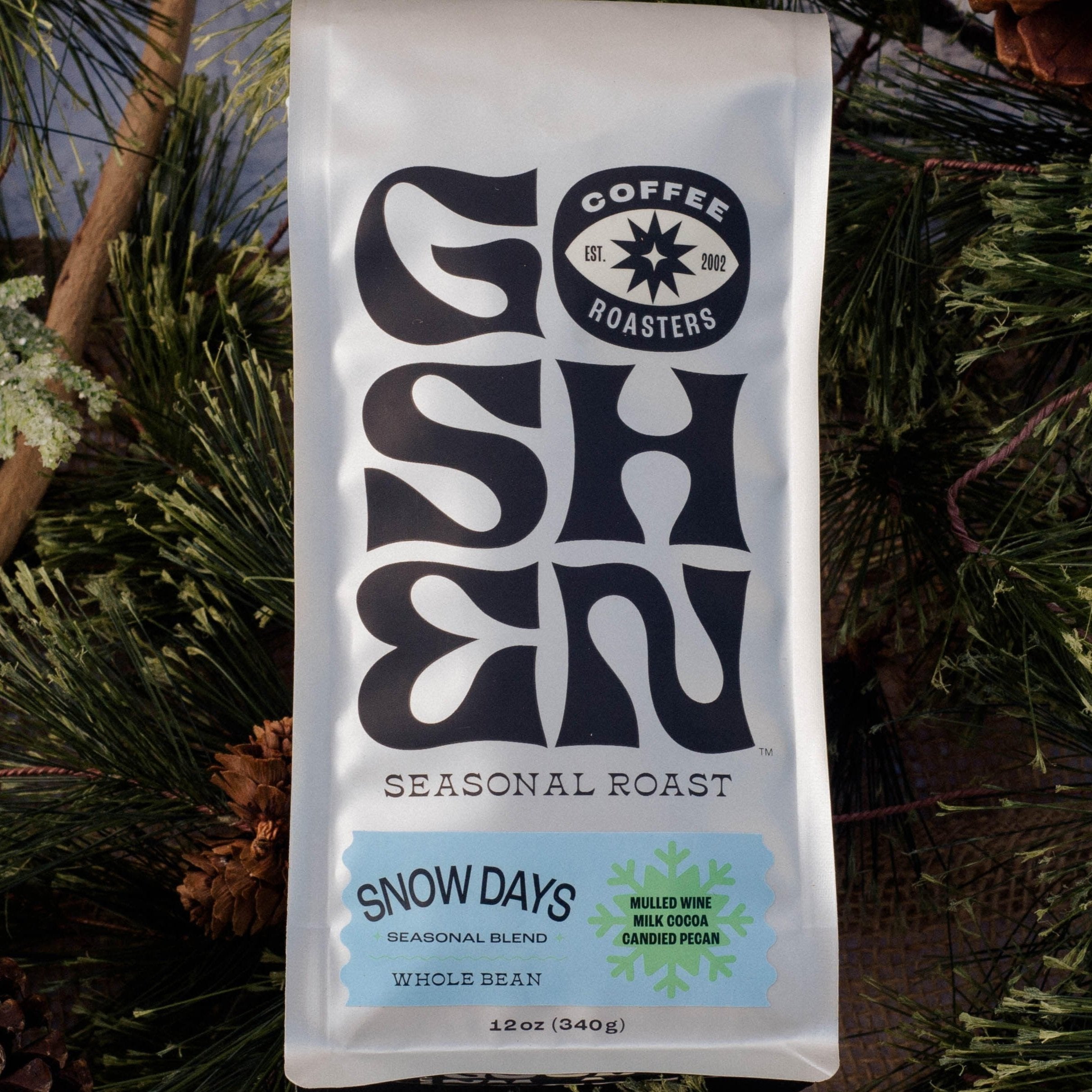 white bag of Goshen Coffee Roasters' "Snow Days" seasonal roast placed amidst a festive arrangement of pine branches, twigs, and pinecones. The bag features bold black lettering and a snowflake design. Flavor notes include "Mulled Wine, Milk Cocoa, Candied Pecan," with the text "Seasonal Blend, Whole Bean" and a weight of 12 oz (340g).

