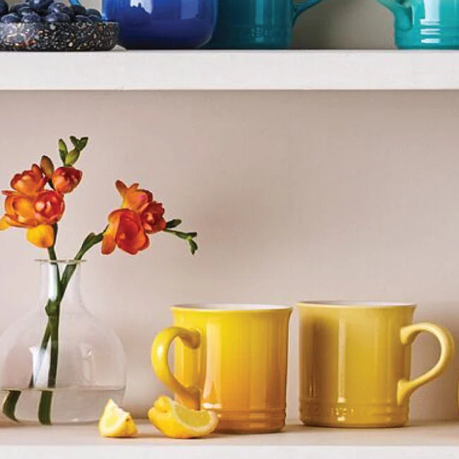 Two yellow mugs on a shelf with a vase of flowers and a lemon.