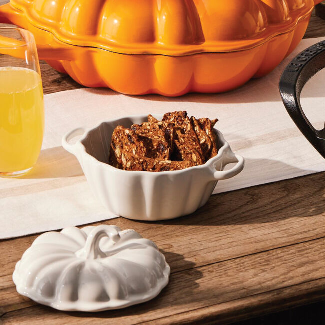 White ceramic pumpkin-shaped lid on a wooden table with a glass of beer and a bowl of cookies.