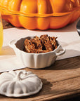 White ceramic pumpkin-shaped lid on a wooden table with a glass of beer and a bowl of cookies.