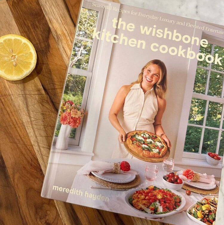 Cookbook titled 'the wishbone kitchen cookbook' by Meredith Hayden on a wooden surface with a lemon slice.