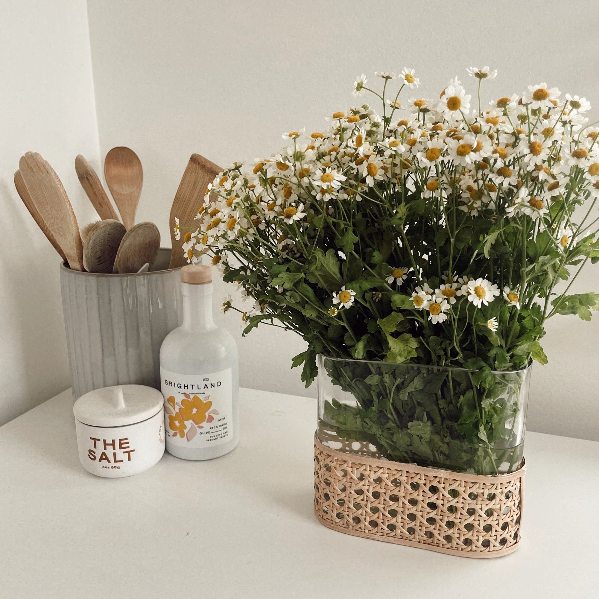 A rattan-wrapped glass vase holding white and yellow daisies next to a utensil jar, a salt cellar, and an olive oil bottle on a white table against a white background