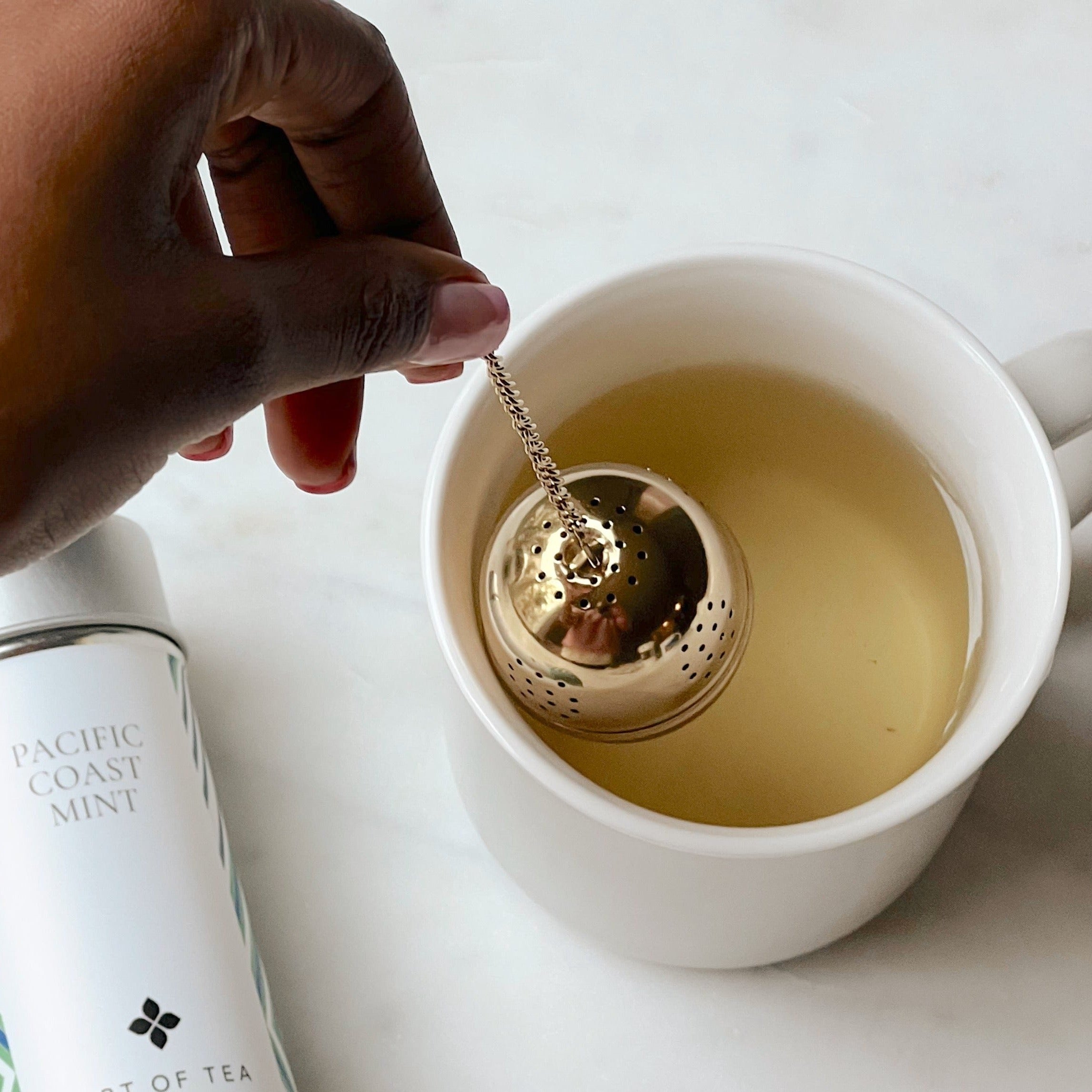A woman's hand dipping a gold acorn tea infuser into a light grey ceramic mug holding hot water. Next to the mug is Art of Tea Pacific Coast Mint tea in a tin.