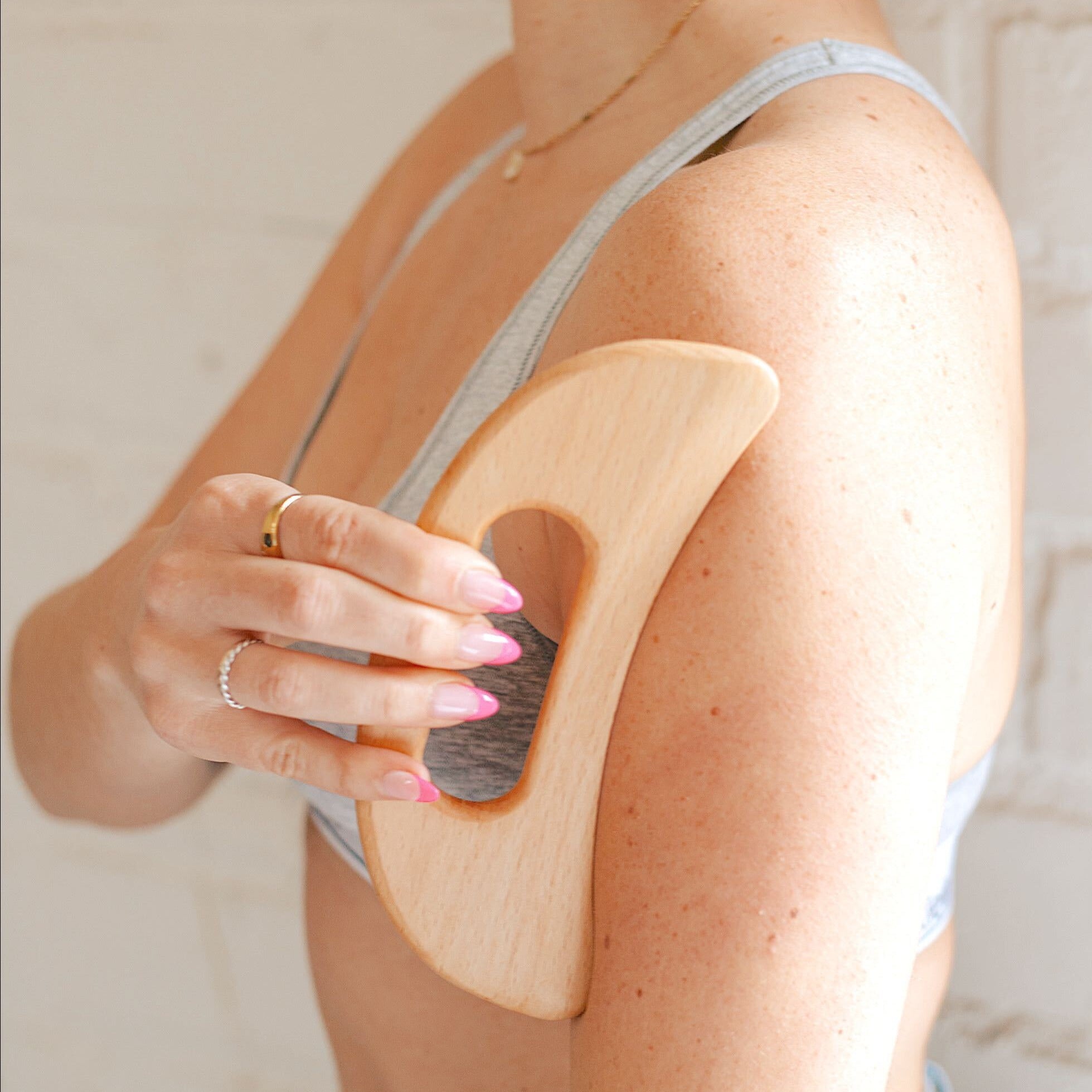 A woman using a wooden lymphatic drainage tool on her left arm while her right hand holds the tool against her skin. She is wearing a gray sports bra standing in front of a white brick background.
