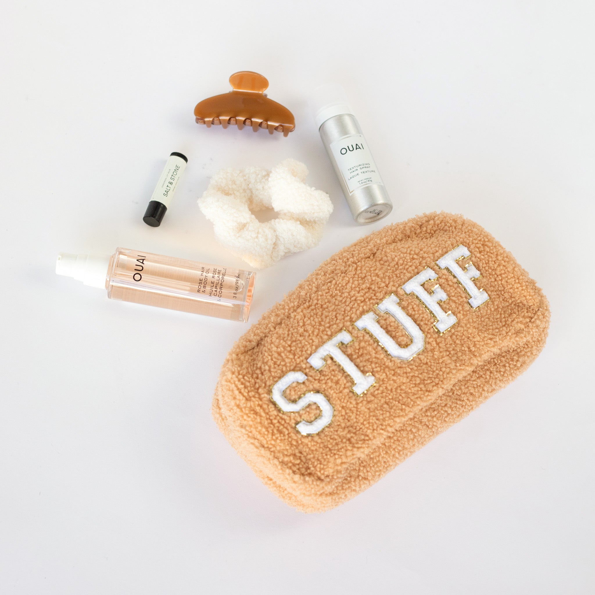 A brown sherpa teddy medium-sized cosmetic bag with gold-trimmed white letters spelling out the word "STUFF" across the length of the bag. Laying on the white background is a bottle of peach colored Ouai hair oil, a Salt + Stone chapstick, a winter white sherpa hair scrunchie, a spray can of Ouai texturizing hair spray, and a caramel colored claw clip.