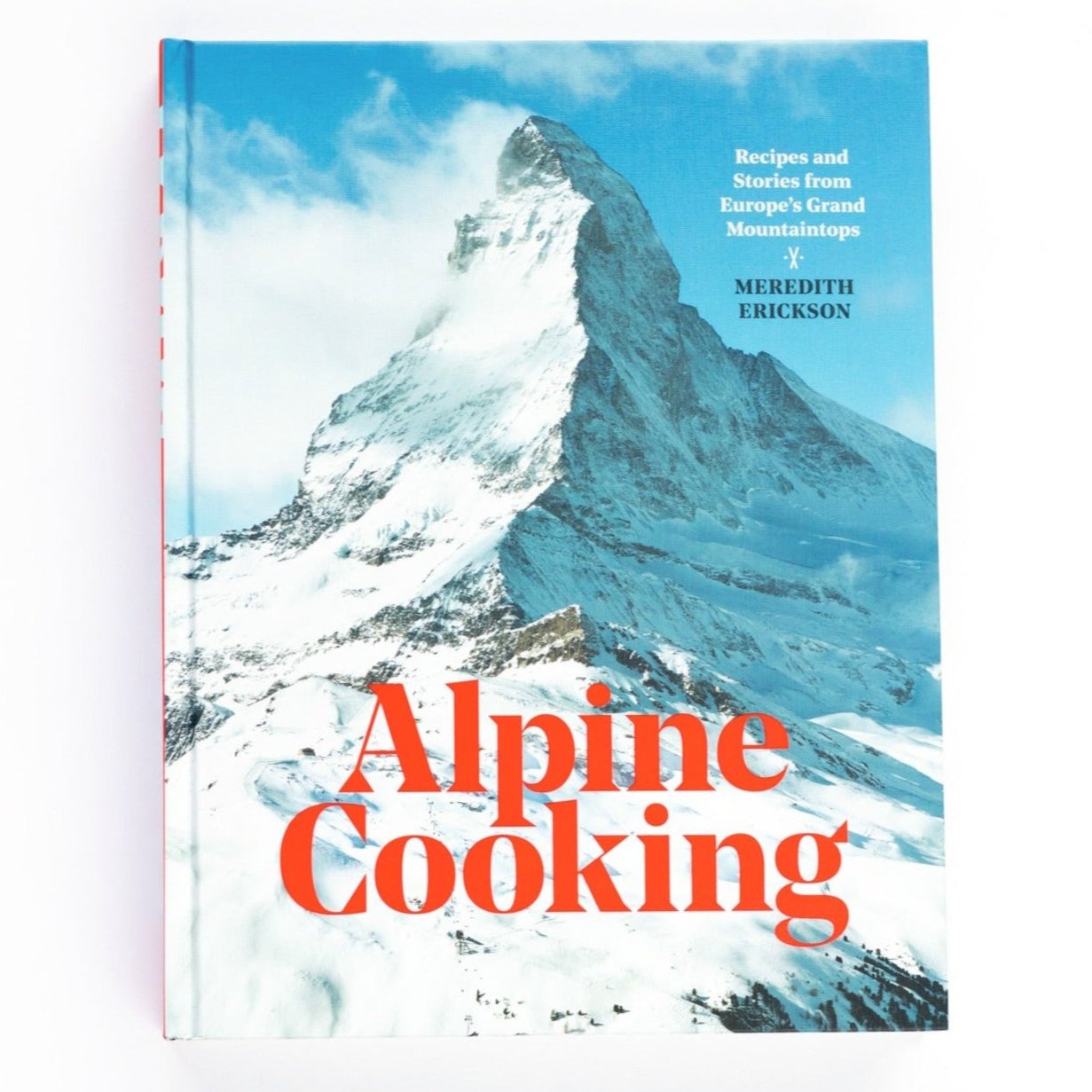 A hardcover book with photo of snow covered mountain peak on cover with red text that reads, "Alpine Cooking" photographed on white background.