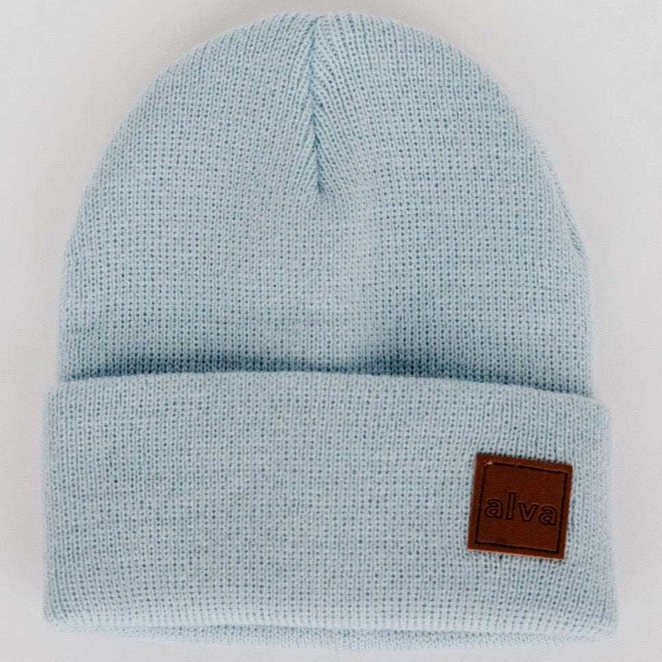 A dusty blue colored ribbed baby beanie with a small square faux leather patch that says "alva" on the bottom right corner. The hat is sitting on a white background