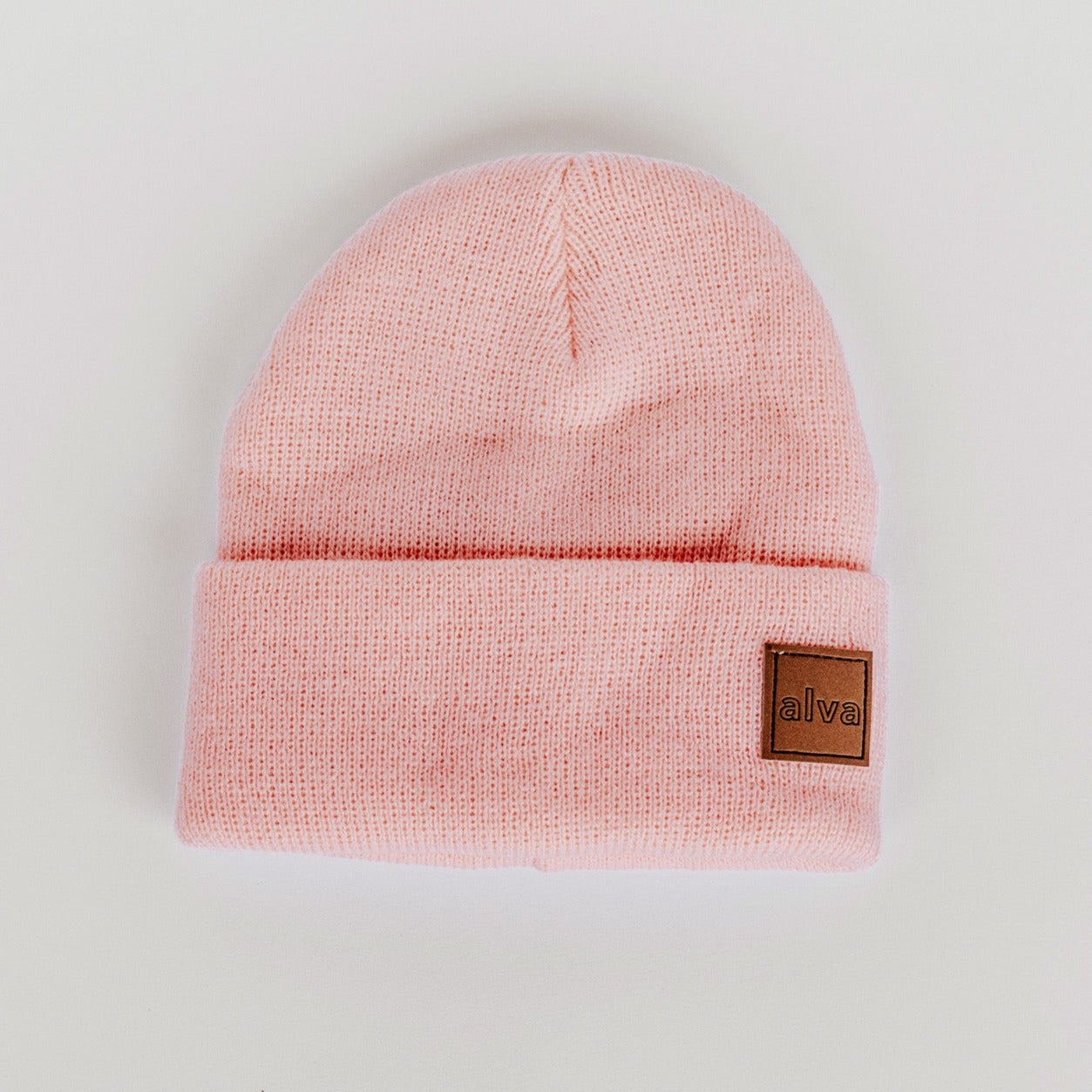 A light pink colored ribbed baby beanie with a small square faux leather patch that says "alva" on the bottom right corner. The hat is sitting on a white background