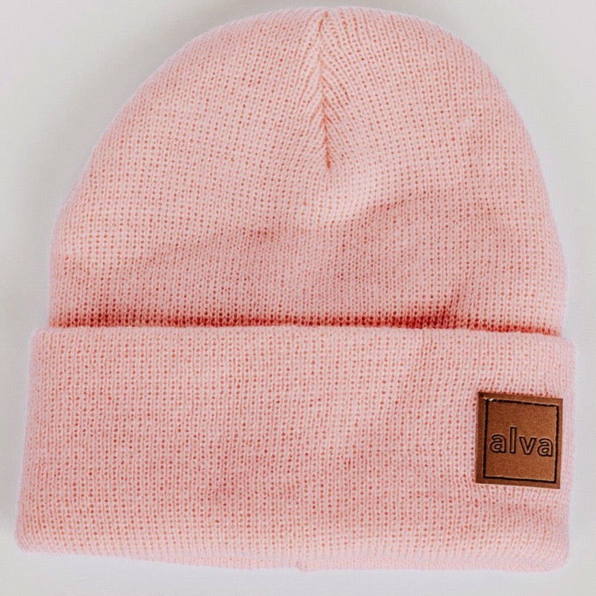A light pink colored ribbed baby beanie with a small square faux leather patch that says "alva" on the bottom right corner. The hat is sitting on a white background