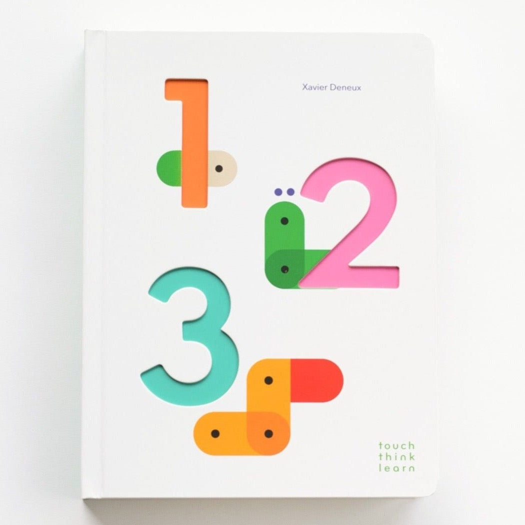 A white board book with cutouts in the shapes of numbers 1, 2 and 3 with colored paper on first page showing through. Text on book reads, "Xavier Deneux touch think learn". Photographed on white background.