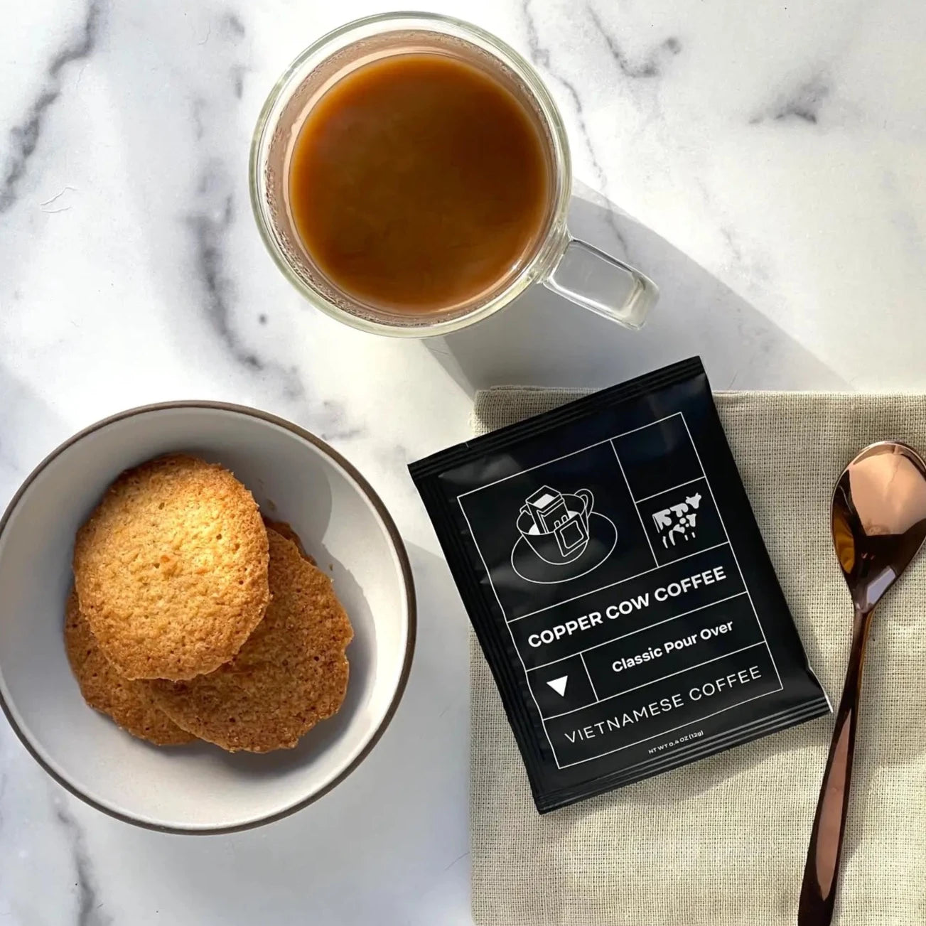 Black packaging for single serving coffee next to a spoon, plate of cookies and mug of coffee.