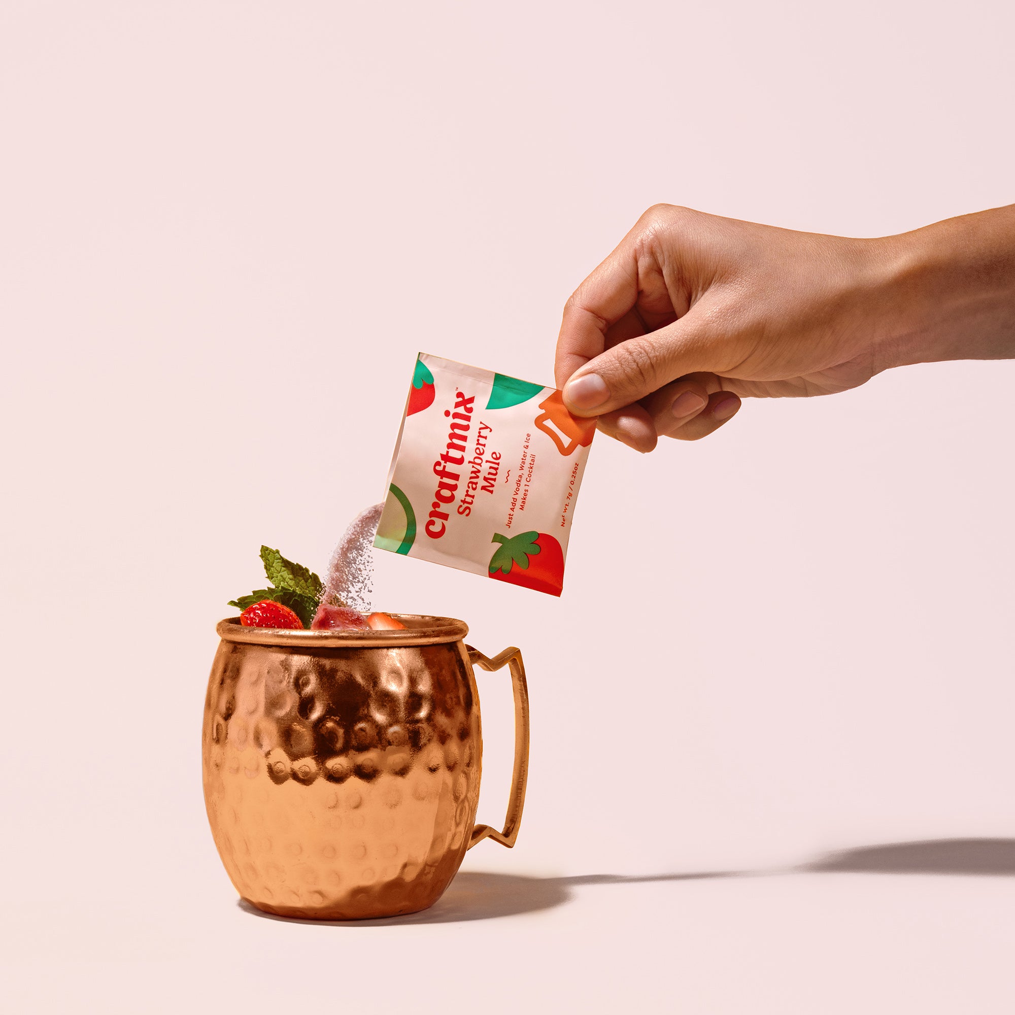 Packet of Craftmix Strawberry Mule being poured into copper Moscow mule mug with strawberry and mint garnish.