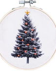 white fabric with black Christmas tree printed on it. Embroidered onto the tree are orange ornaments and a gold star tree topper. Embroidery hoop is light brown with silver metal clasp