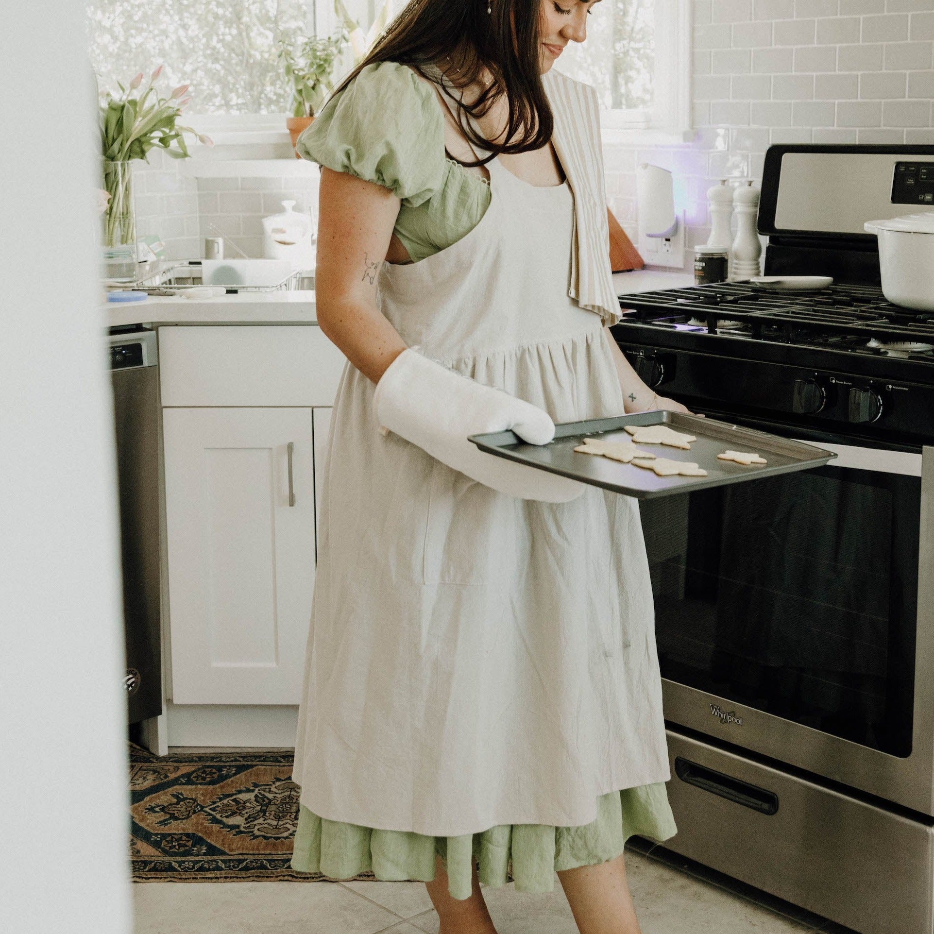 A woman wearing a green dress with an oatmeal colored kitchen apron. She is holding a tray of sugar cookies in a kitchen.