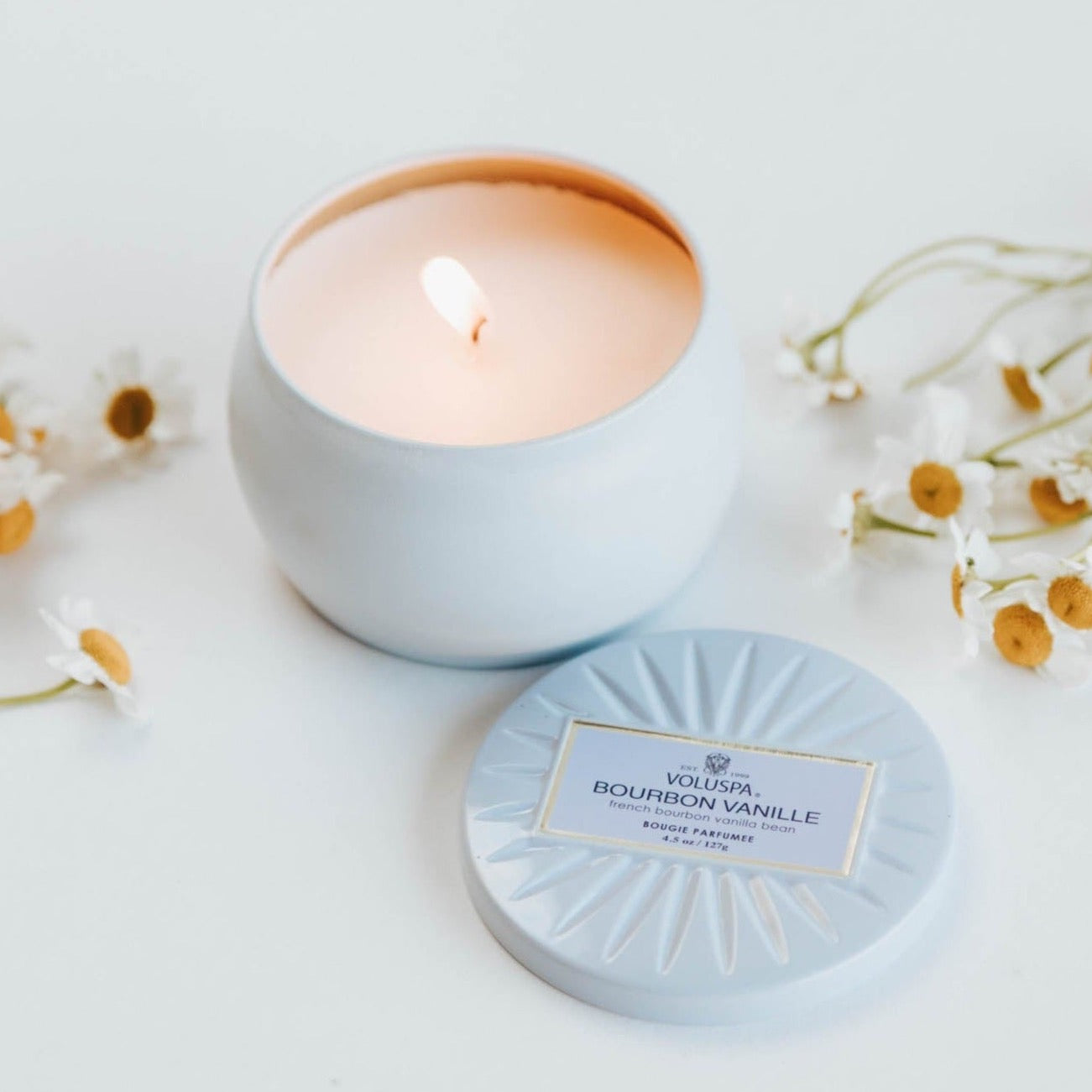 Lit Bourbon Vanille tin candle on white background with chamomile flowers surrounding.