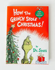 Hardcover of "How the Grinch Stole Christmas" 2021 book against a white background. The cover is red on top, white on the bottom with a cartoon decorated Christmas tree on the left, a cartoon grinch in the middle, and one blue and one red present on the right.