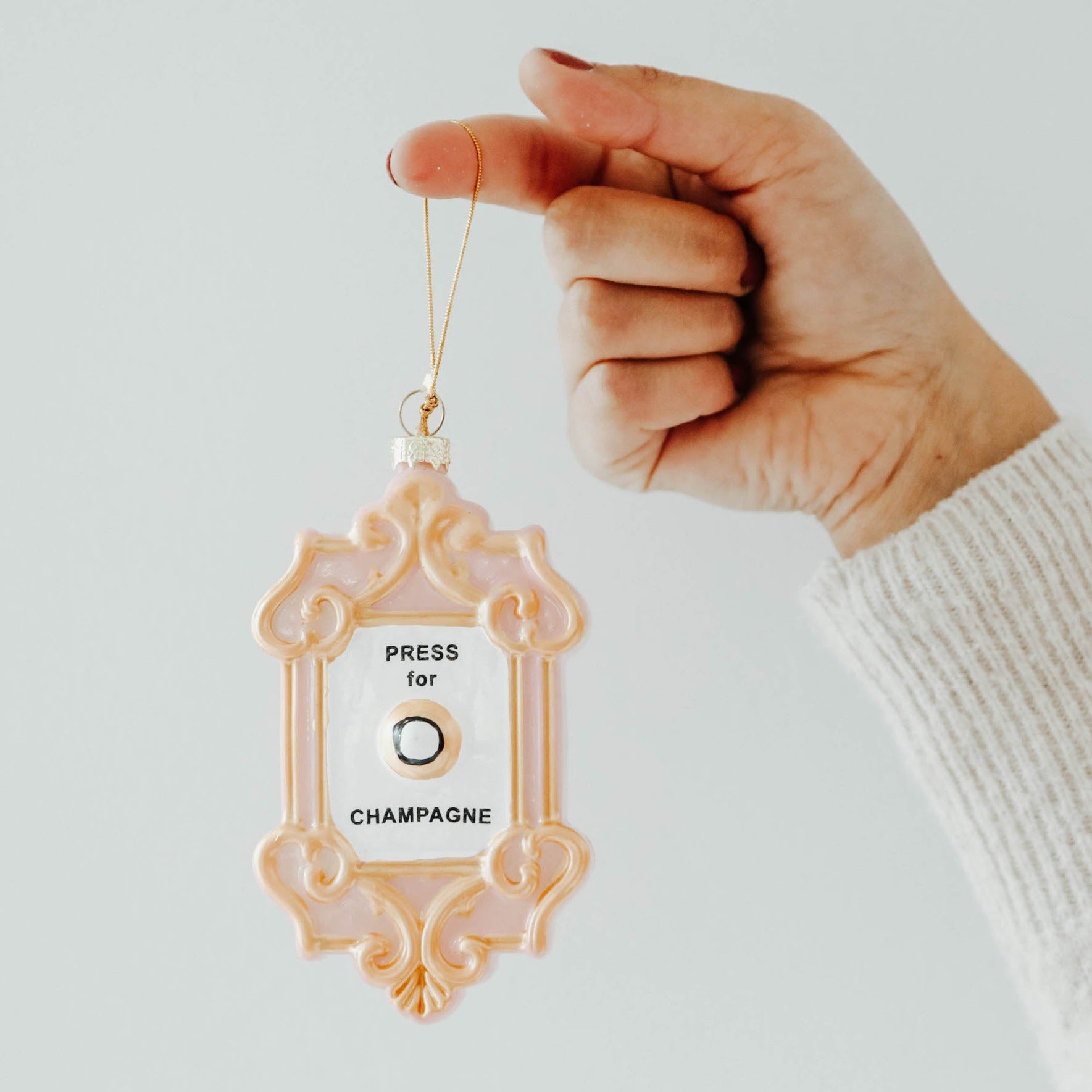 A woman's hand holding a pink "Press for Champagne" button ornament against a white background. 