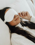 A girl with long wavy brunette hair wearing a white sweater laying on a white bed with an ivory silk eye mask over her eyes.