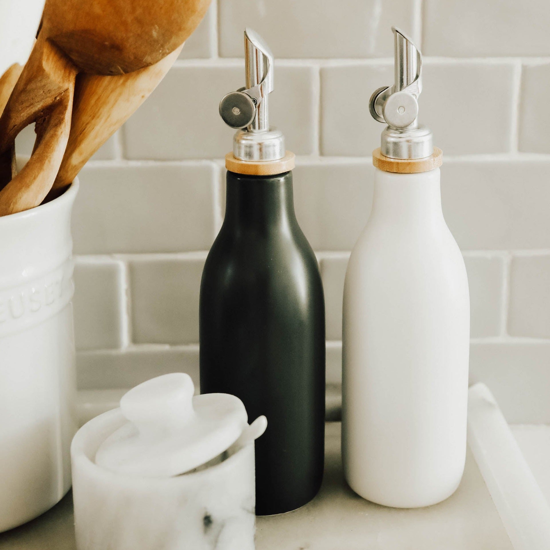 A gray oil cruet to the left of a white oil cruet sitting on a white marble tray behind a white marble salt cellar.