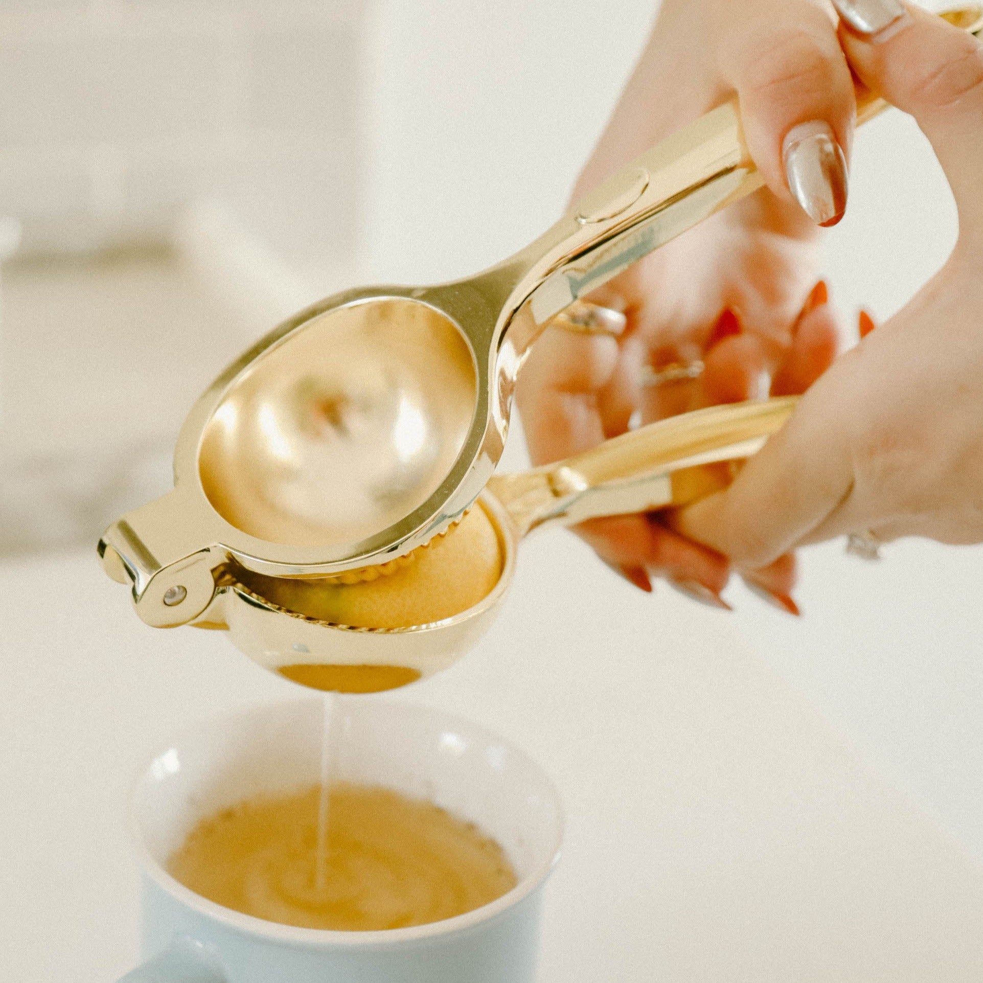 A woman's hands using a gold metal citrus press to squeeze half a lemon into a blue mug holding tea. The background is white and blurred out.