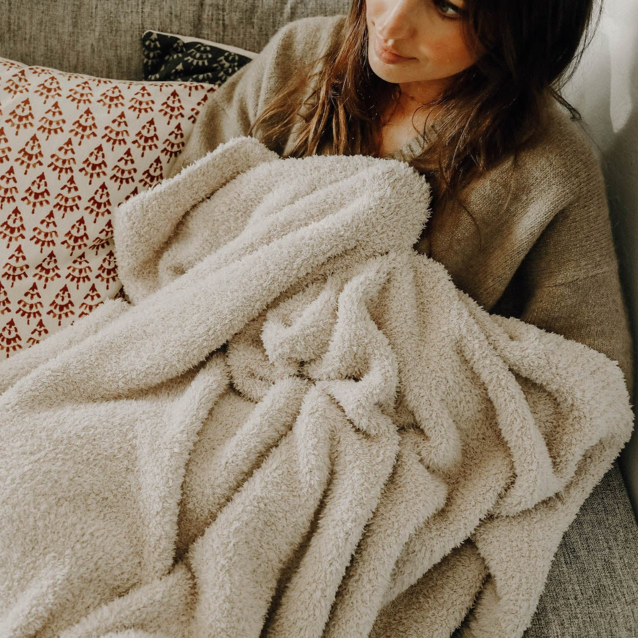 A brunette girl laying on a couch with an oatmeal plush blanket wrapped around her.