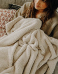 A brunette girl laying on a couch with an oatmeal plush blanket wrapped around her.