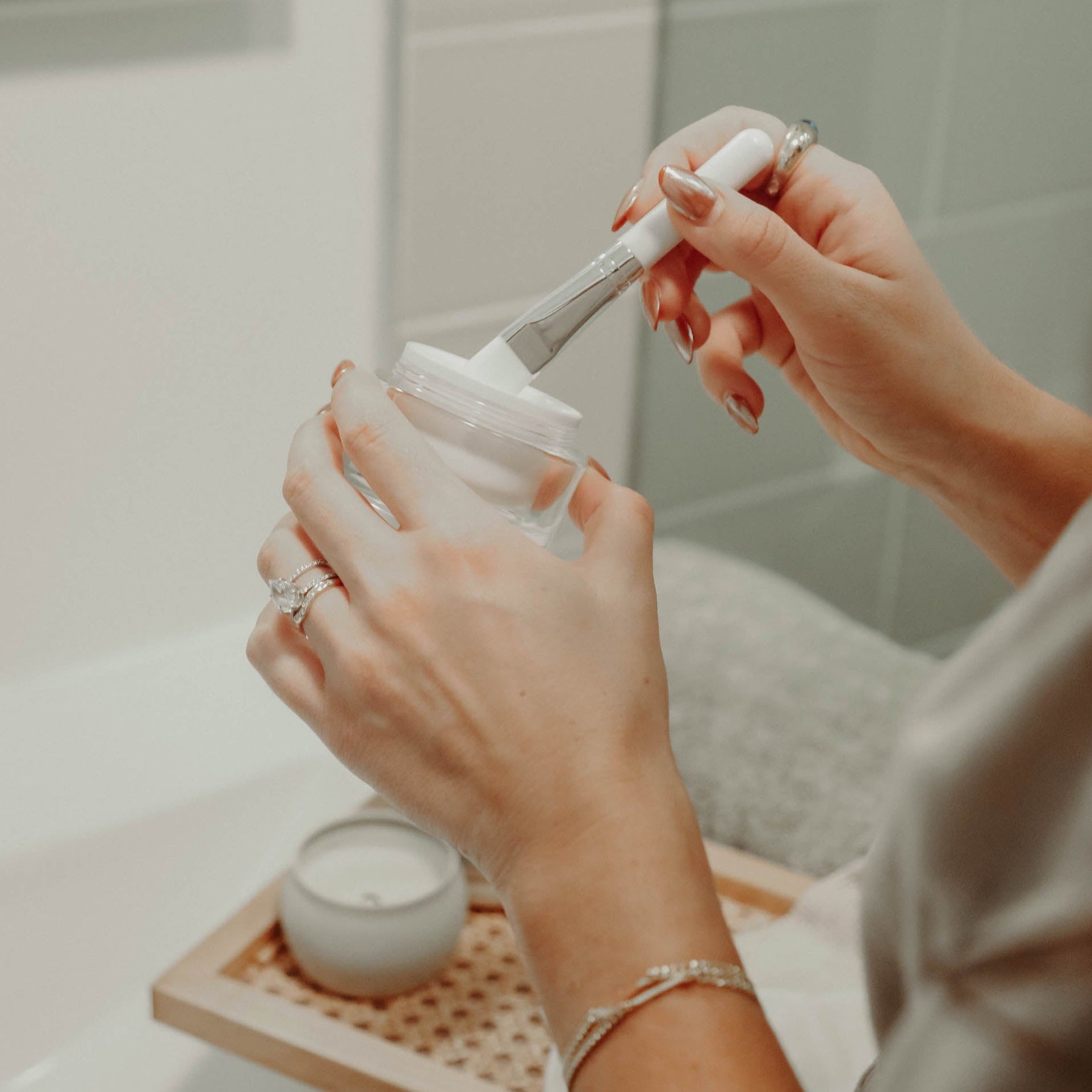 Woman's hands dipping a face mask applicator brush into a pot of face lotion.